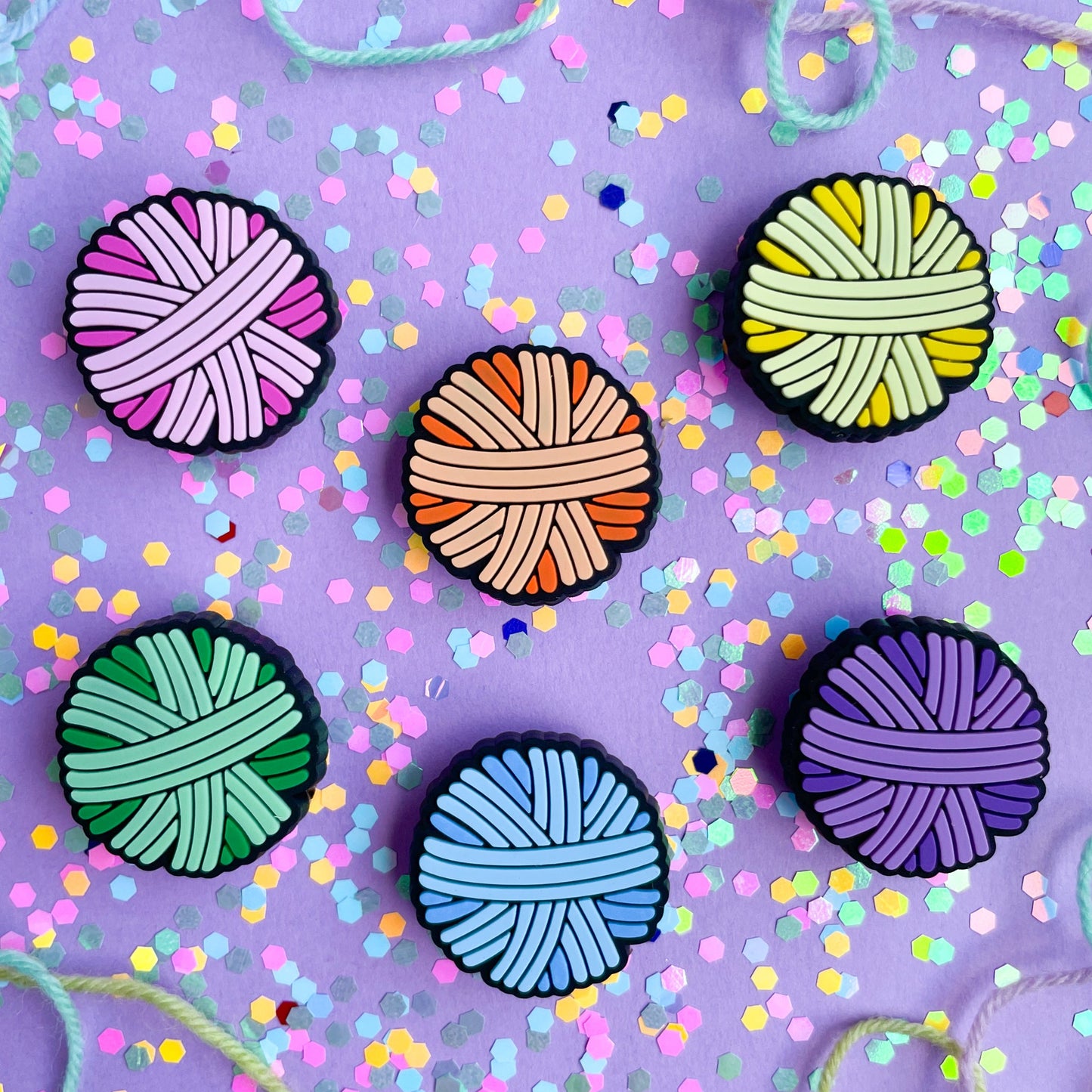 PVC charms shaped like yarn balls in pink, orange, yellow, mint, blue, and purple on a lavender background covered in confetti.