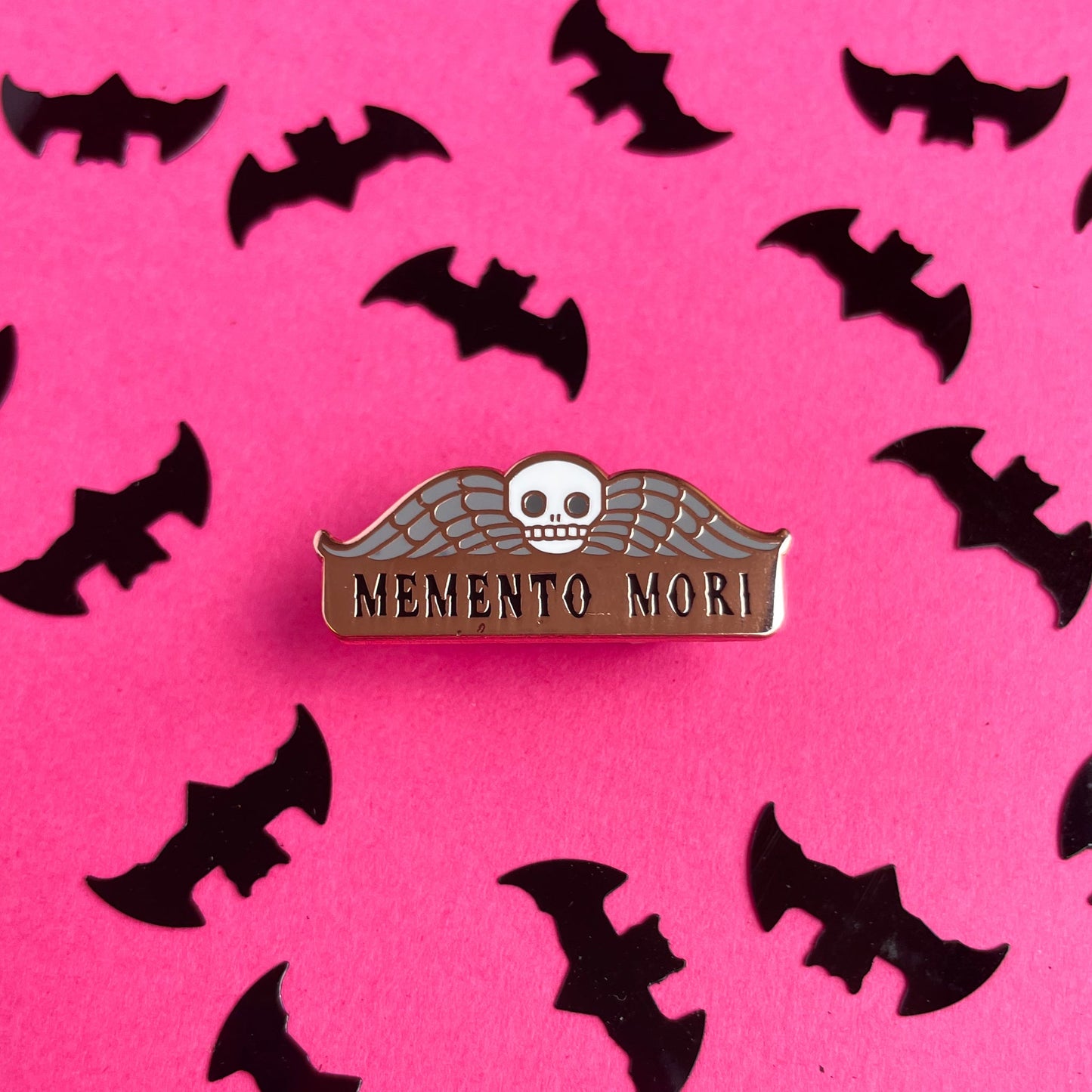 An enamel pin that reads "Memento Mori" with a skull and wings on it on a hot pink background surrounded by bat confetti. 