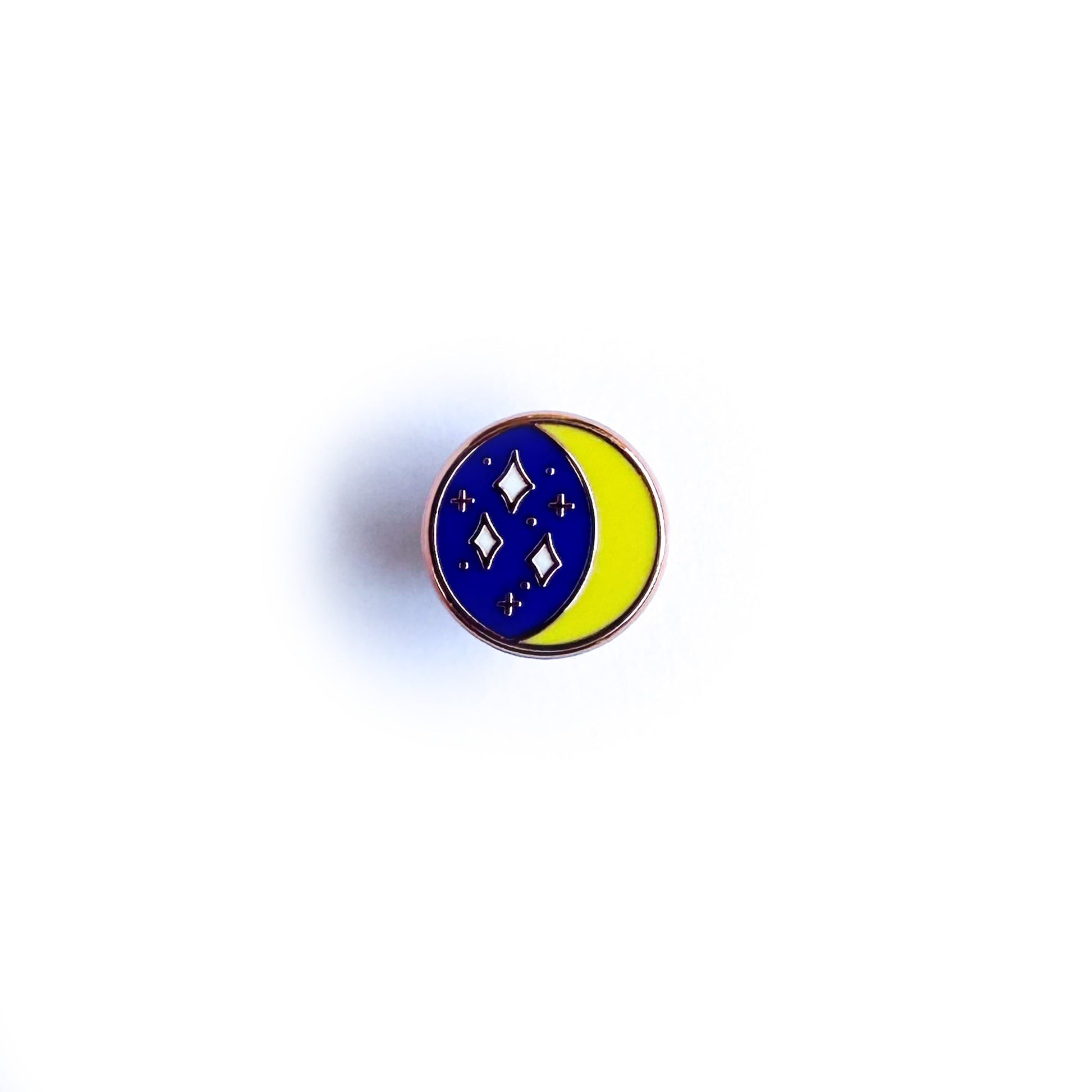 A circular enamel pin with an image of a crescent moon and a starry sky inside it. 