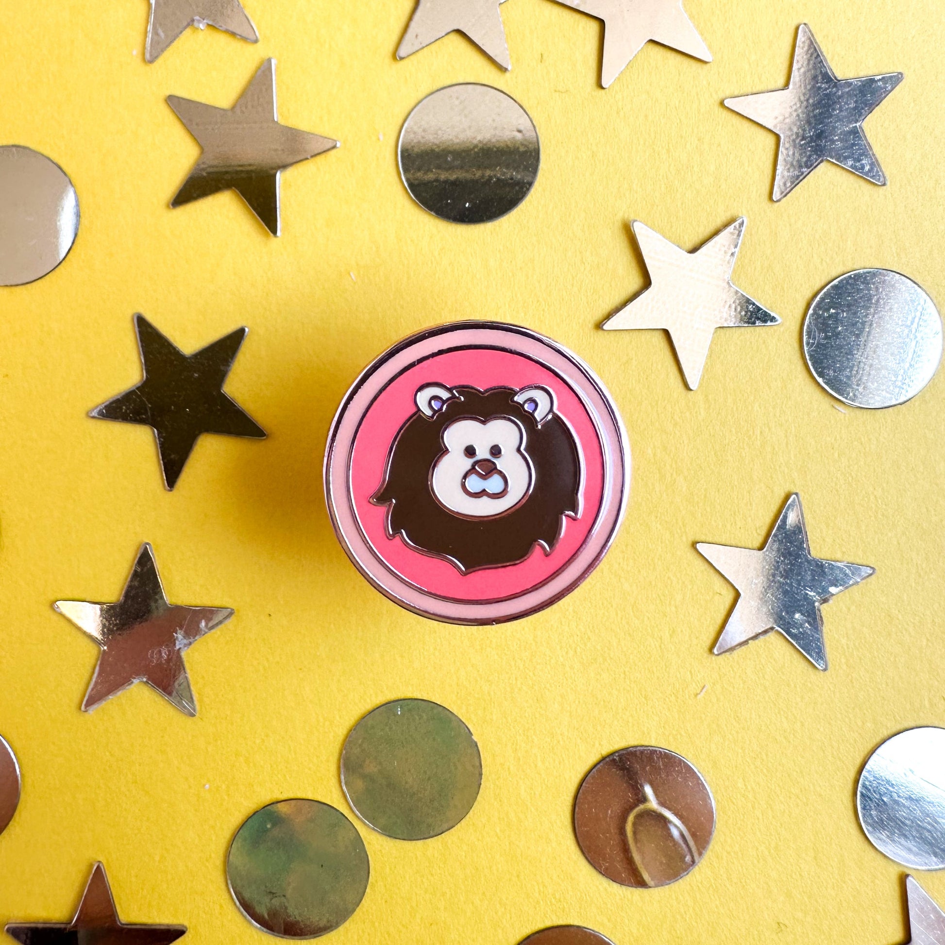 A circle enamel pin with a cute Lion image in it to represent the Leo zodiac sign. The pin is on a yellow background covered in gold star and circle confetti. 