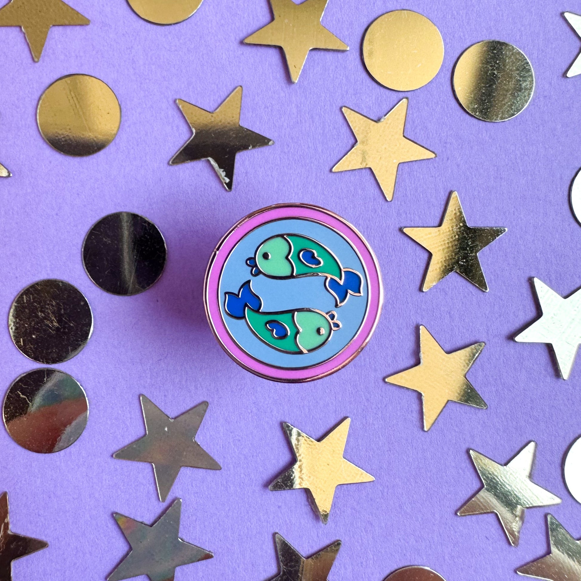 A pin with cute fish swimming in opposite directions to represent Pisces. The pin is on a purple paper background with gold star and circle confetti surrounding it. 
